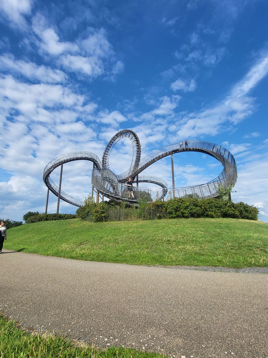 Een sculptuur van stalen trappen die de vorm van een achtbaan nabootsen, gelegen op een groene heuvel onder een blauwe lucht met witte wolken. Er zijn mensen zichtbaar op de trappen en een persoon loopt op het pad links van de heuvel.