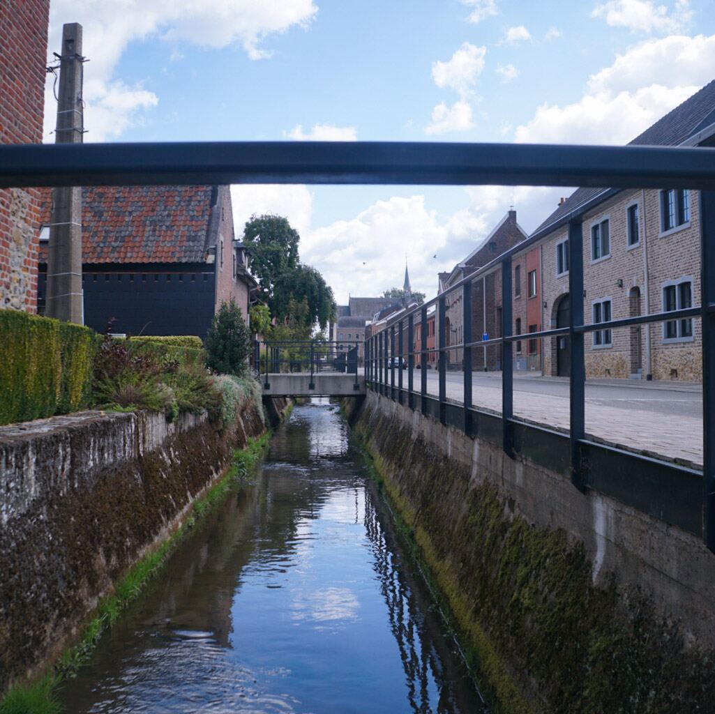 Een smal kanaal met water stroomt door een stedelijke omgeving, geflankeerd door stenen muren met mos en aan weerszijden huizen. Op de achtergrond is een kerktoren zichtbaar en de lucht is deels bewolkt.