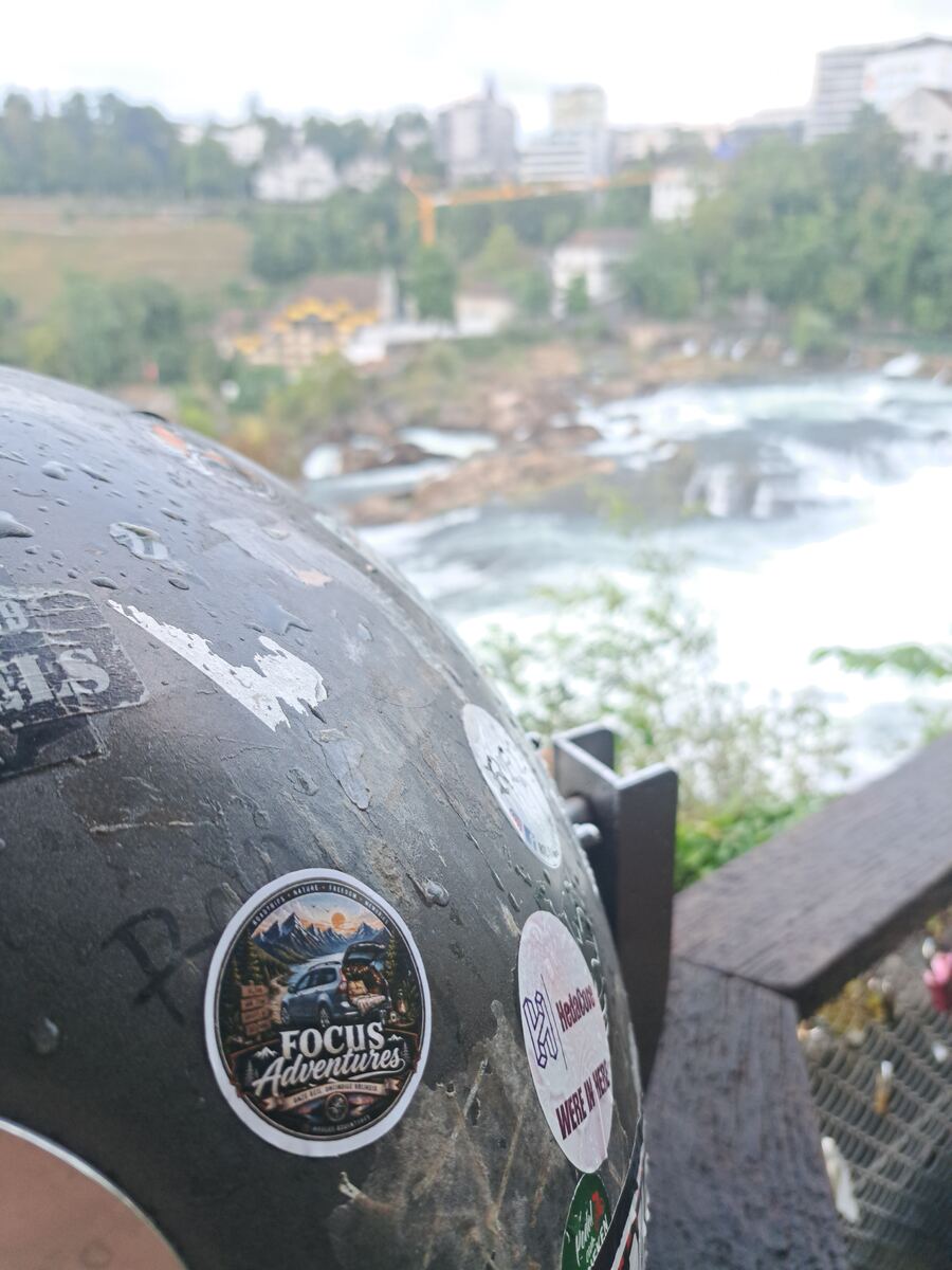 De voorgrond toont een donker, met stickers beplakt en met waterdruppels bedekt oppervlak, mogelijk een helm of uitrustingsstuk. Een sticker met 'FOCUS Adventures' en een auto in een berglandschap is duidelijk zichtbaar. De achtergrond, onscherp, toont een brede rivier met stroomversnellingen en rotsen in het water. Aan de overkant van de rivier zijn groene begroeiing, gebouwen en enkele grotere constructies te zien, waaronder een kraan. Rechts is een deel van een houten reling zichtbaar.