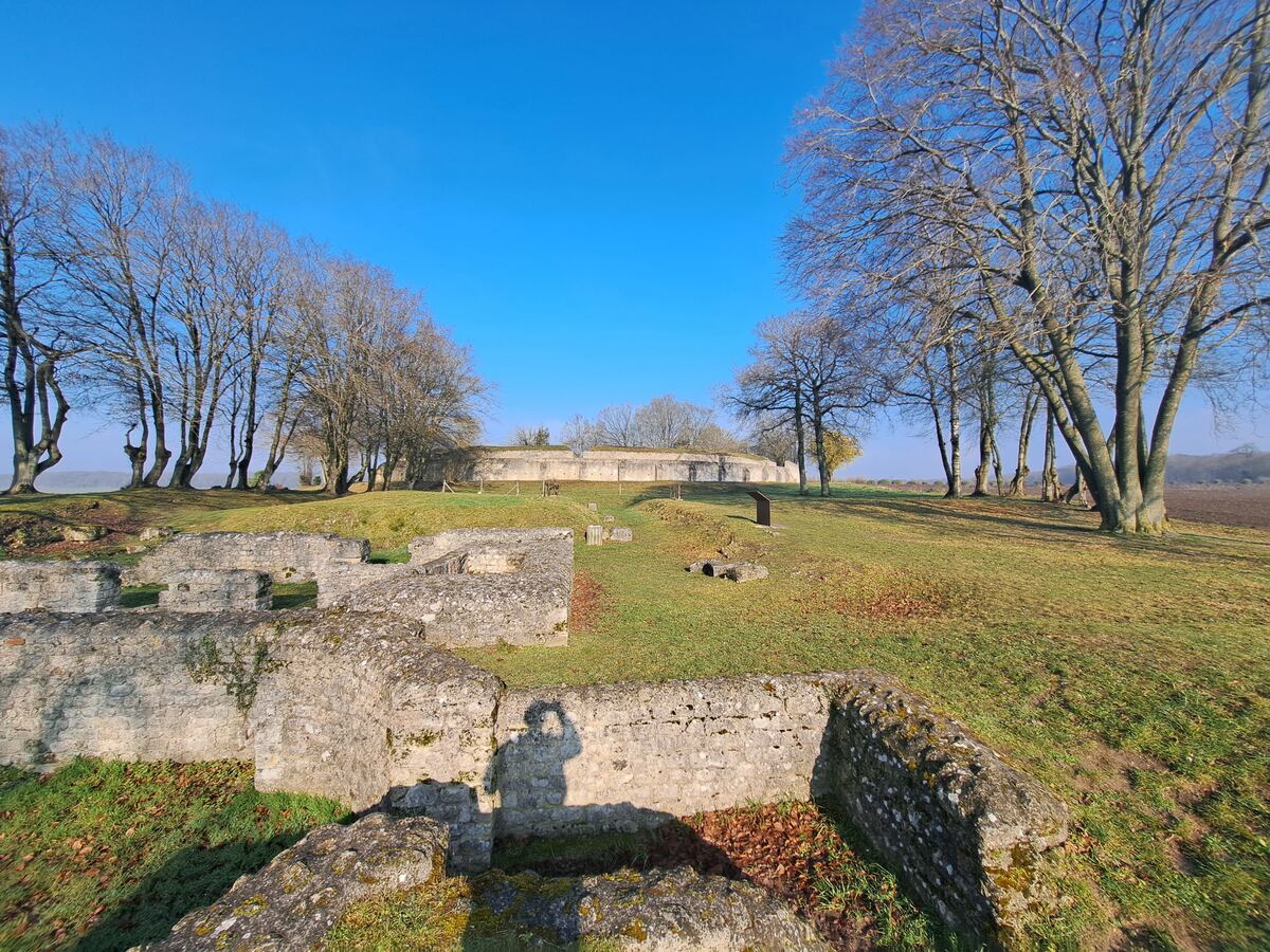 De foto toont de stenen fundamenten van een oud bouwwerk op een zonnige dag. Op de achtergrond is een ronde, met gras begroeide aarden wal te zien, omzoomd door kale bomen. De zon werpt lange schaduwen, waaronder die van een persoon die een foto maakt.