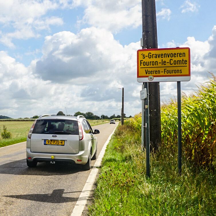 Een zilverkleurige Ford Focus stationwagen staat geparkeerd langs een landelijke weg, met een geel plaatsnaambord voor 's-Gravenvoeren, Fouron-le-Comte en Voeren-Fourons ernaast. De weg loopt door een groen landschap met akkers en bomen onder een deels bewolkte lucht.
