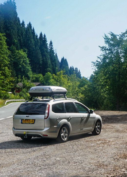 Een zilverkleurige Ford Focus stationwagen met een dakkoffer staat geparkeerd op een onverharde strook naast een bochtige weg. De auto is van achteren en licht van opzij te zien. Op de achtergrond zijn dichtbegroeide, groene bergen onder een helderblauwe lucht zichtbaar.