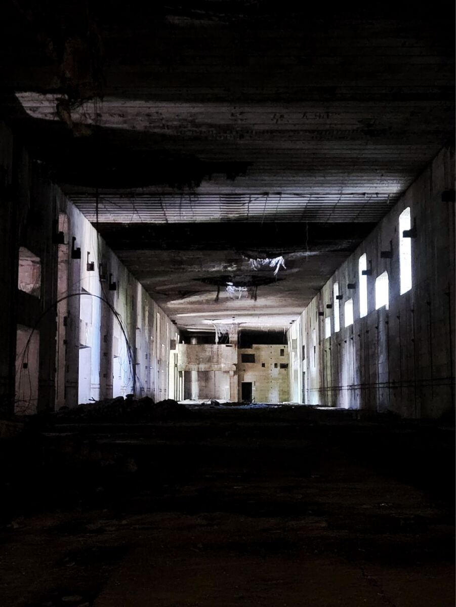 Een lange, donkere gang in een betonnen bunker met licht dat door ramen aan de zijkanten valt. De gang is leeg en de muren zijn ruw en onafgewerkt. Er zijn enkele kabels en puin zichtbaar.