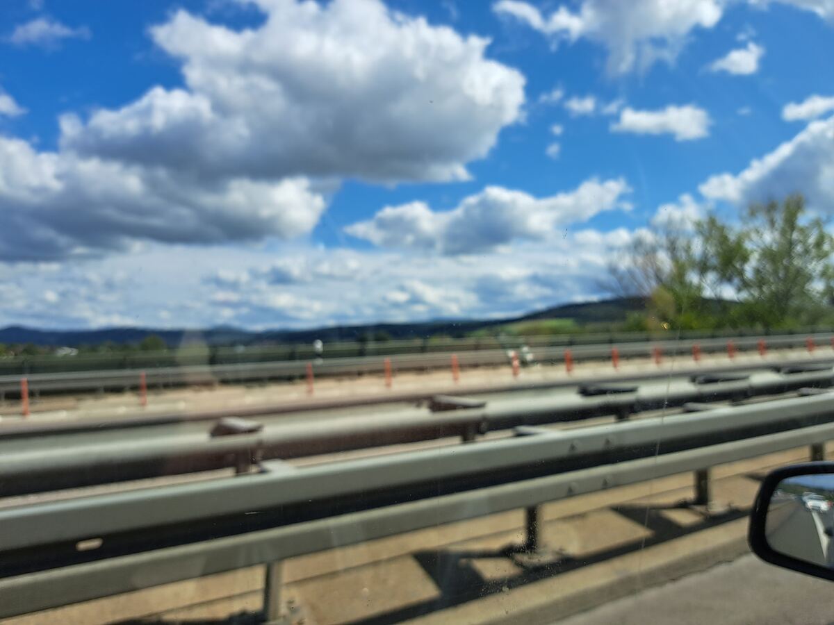 De foto toont een onscherp uitzicht vanuit een rijdende auto op een snelweg. Op de voorgrond is een vangrail te zien. Daarachter liggen meerdere rijstroken van een snelweg, met aan de zijkant oranje paaltjes die mogelijk duiden op werkzaamheden of markering. In de verte zijn glooiende, beboste heuvels zichtbaar onder een helderblauwe lucht met verspreide witte stapelwolken. Een deel van de buitenspiegel is rechtsonder in beeld.