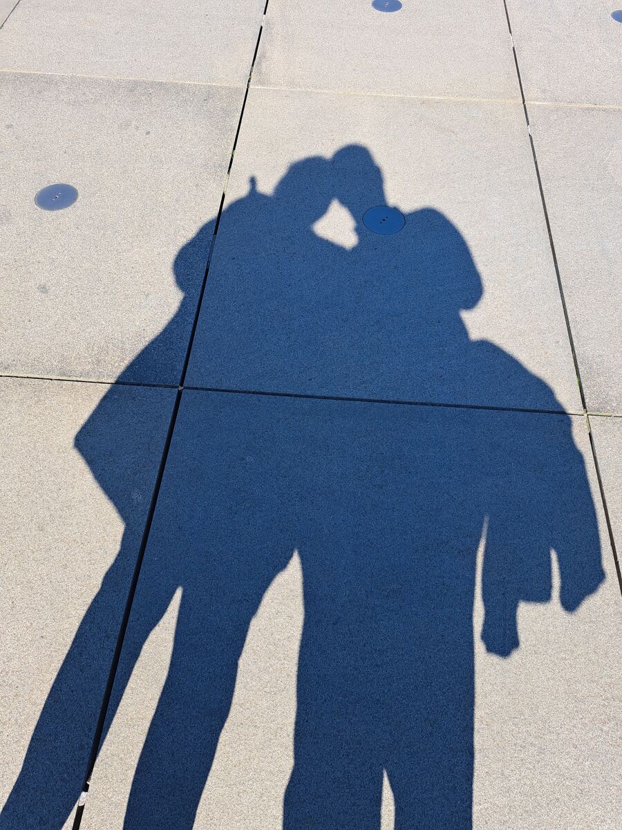De foto toont de schaduwen van twee personen op een lichtgekleurde tegelbestrating. De schaduwen zijn scherp afgetekend, wat duidt op fel zonlicht. Eén van de schaduwen lijkt een arm omhoog te houden, mogelijk om een foto te maken. Er zijn ook enkele donkere, ronde elementen in de bestrating zichtbaar.