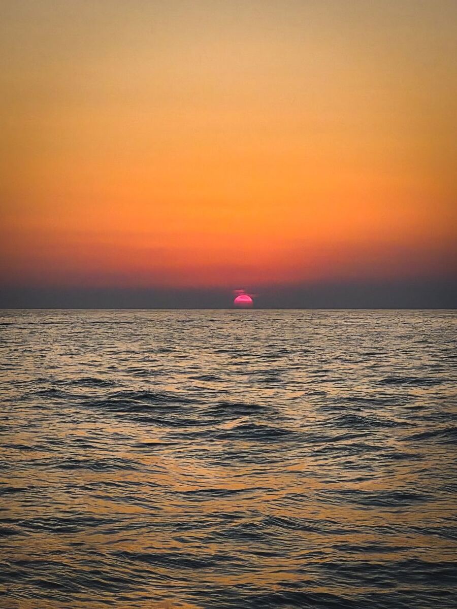 Een foto van een zonsondergang boven een kalme zee. De zon, die als een helder roze-oranje stip aan de horizon verschijnt, werpt een warme gloed over de lucht die van geel-oranje bovenaan naar dieper oranje en roze bij de horizon verloopt. Het wateroppervlak is donker met kleine golven die de kleuren van de lucht reflecteren.