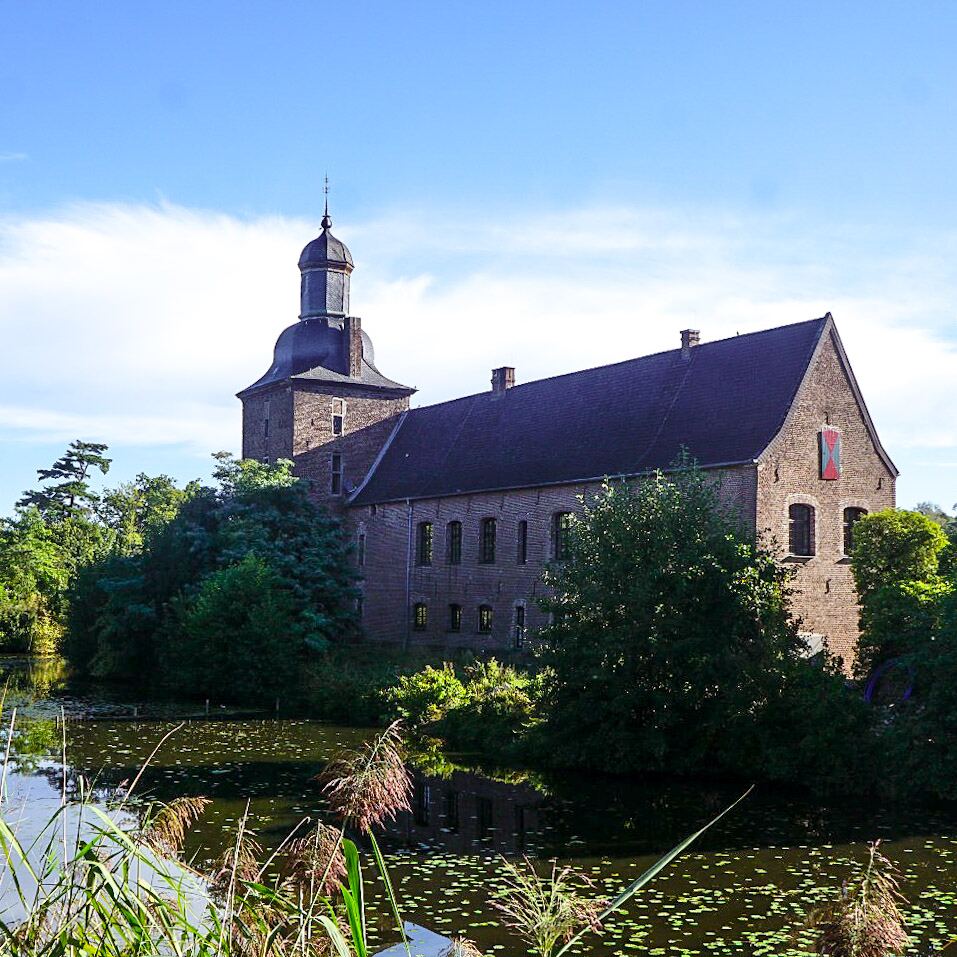 Een bakstenen kasteel met een toren en een donker dak, omringd door water met waterlelies en groene begroeiing onder een blauwe lucht met enkele wolken.