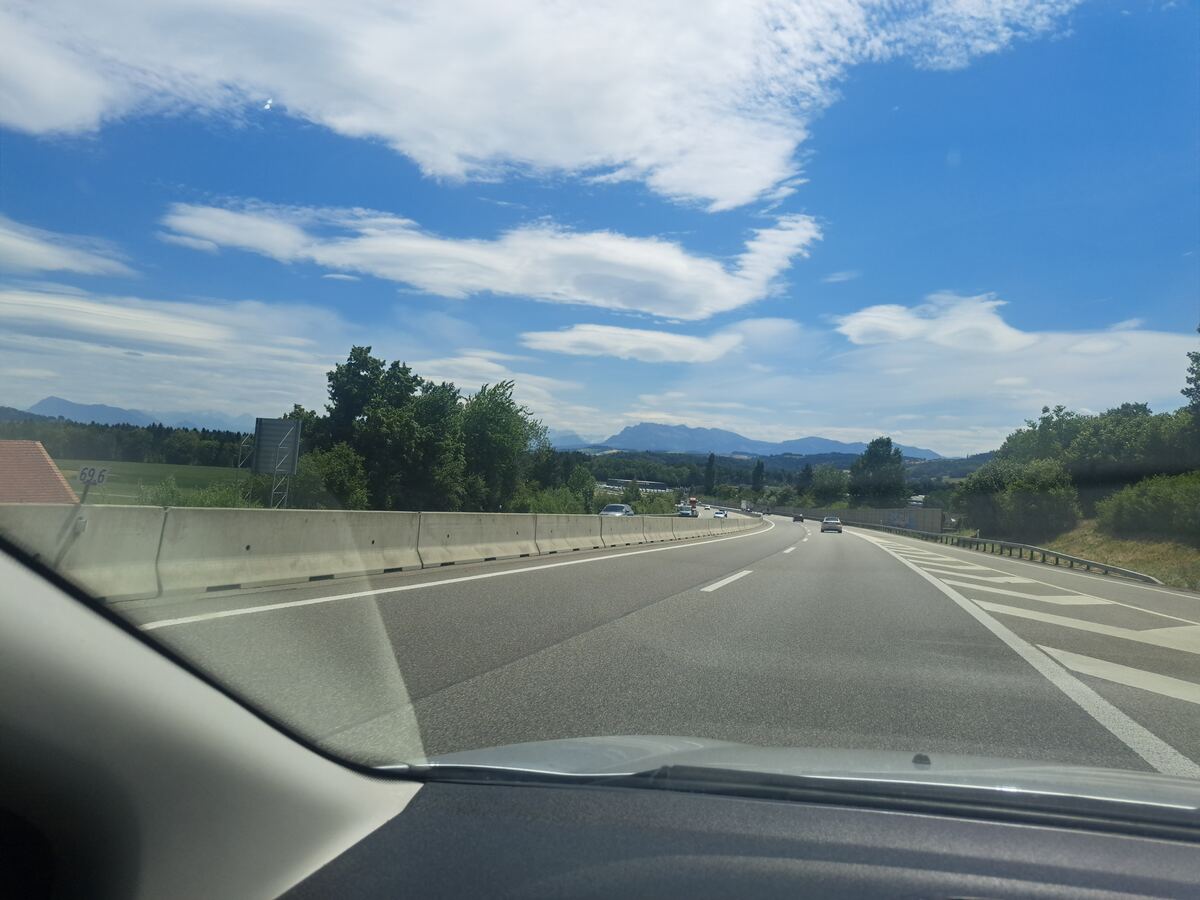 De foto toont een uitzicht vanuit een voertuig op een meerbaanssnelweg die zich uitstrekt naar de horizon onder een deels bewolkte blauwe lucht. Aan de linkerzijde van de weg bevindt zich een betonnen vangrail, daarachter groene vegetatie en bomen, met in de verte een rood pannendak van een gebouw en een wit bord met het nummer "69.6". Rechts van de weg is eveneens dichte begroeiing zichtbaar. In de verre achtergrond tekenen zich bergen af. Er rijden enkele auto's op de snelweg in dezelfde richt
