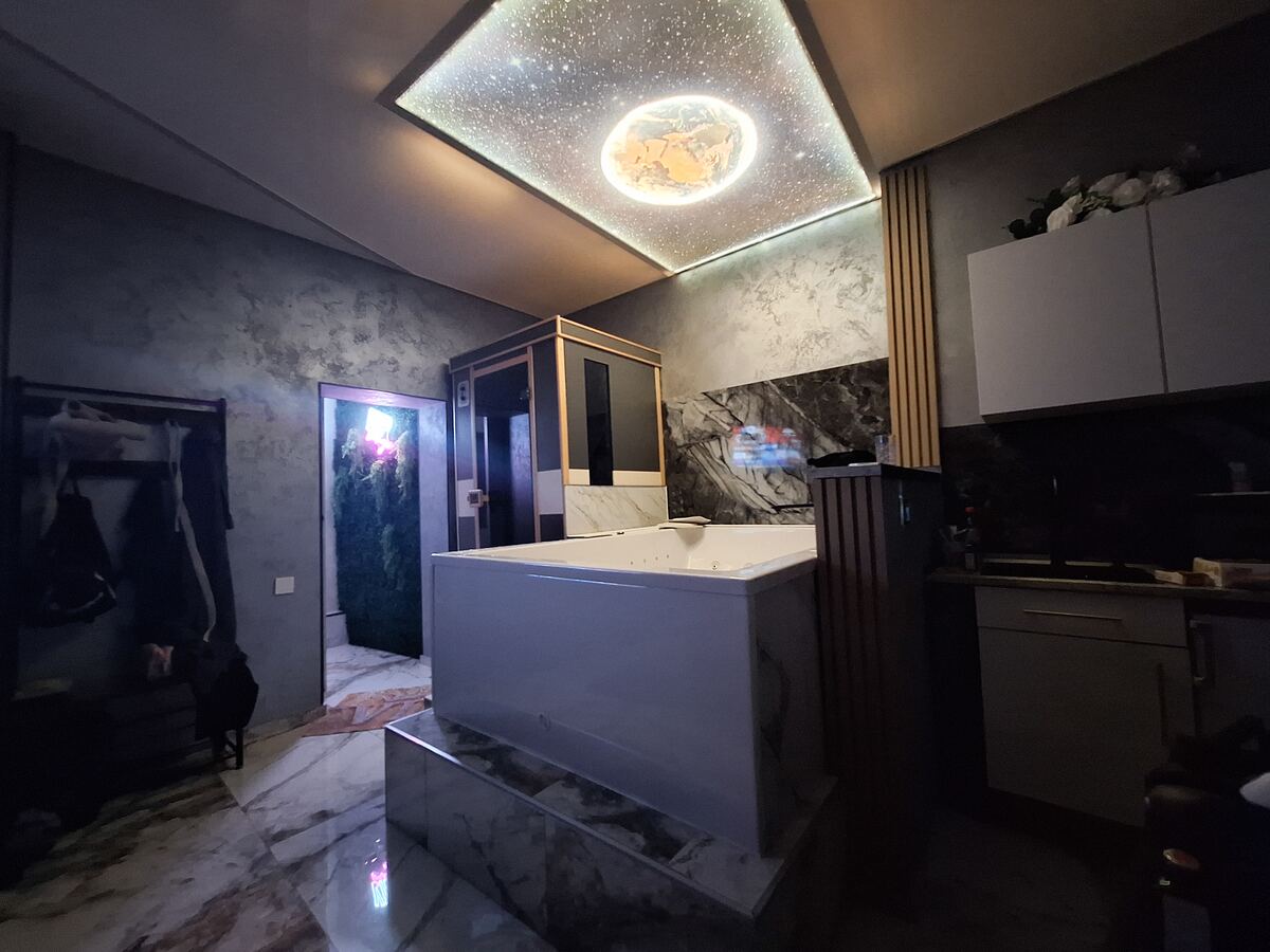 Een sfeervolle hotelkamer of appartement met een groot wit bubbelbad op een verhoging. Boven het bad is een plafond met sterrenhemel en een verlichte bol die op een planeet lijkt. Er is ook een sauna of stoomcabine zichtbaar en een open deur naar een ruimte met groene wanddecoratie. De muren hebben een betonlook en er is een donkere keukenhoek met witte kasten.