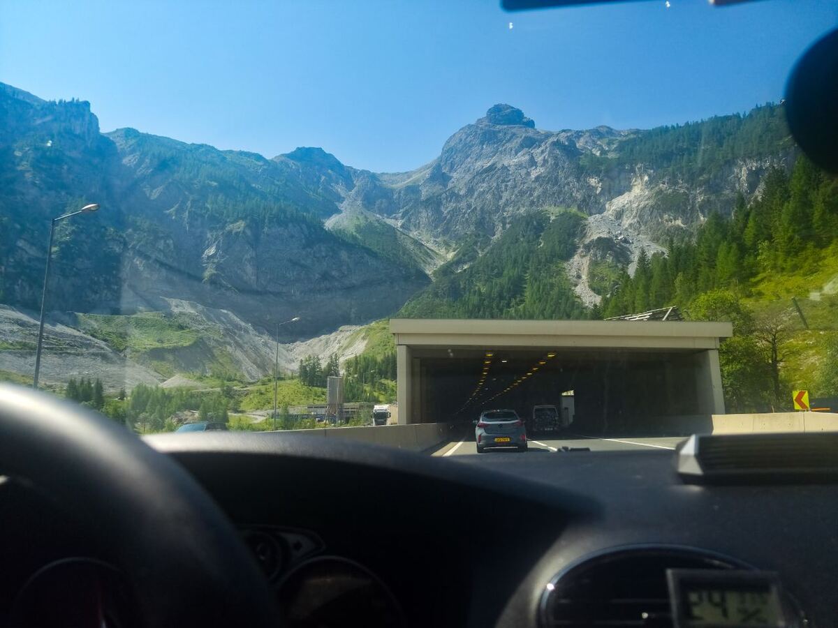 Een foto vanuit de auto op een snelweg die naar een tunnel leidt, met op de achtergrond hoge, deels beboste bergen onder een helderblauwe lucht. De voorgrond toont een deel van het stuur en het dashboard van de auto.