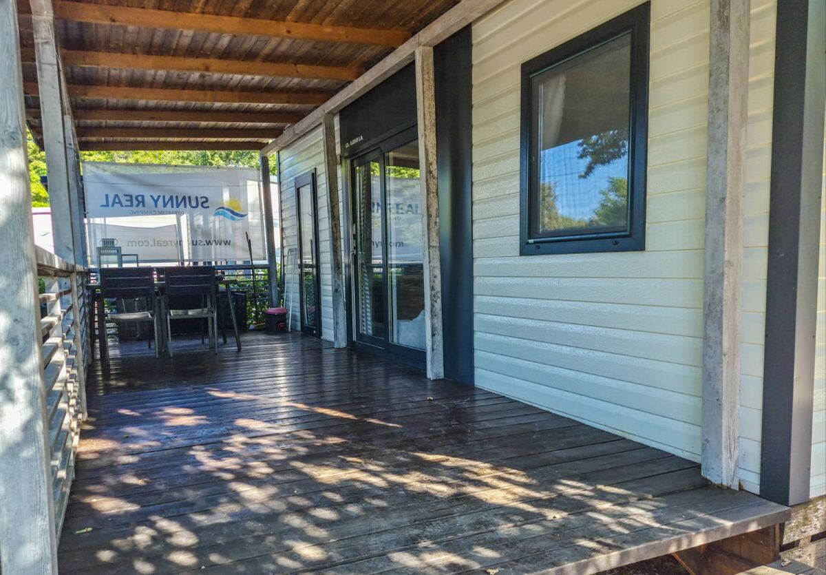 Een houten terras met een overkapping, grenzend aan een stacaravan. Op het terras staan een tafel met stoelen en op de achtergrond is een spandoek met de tekst 'SUNNY REAL' en 'www.sunnyreal.com' te zien. De zon schijnt en werpt schaduwen op de houten planken van het terras.