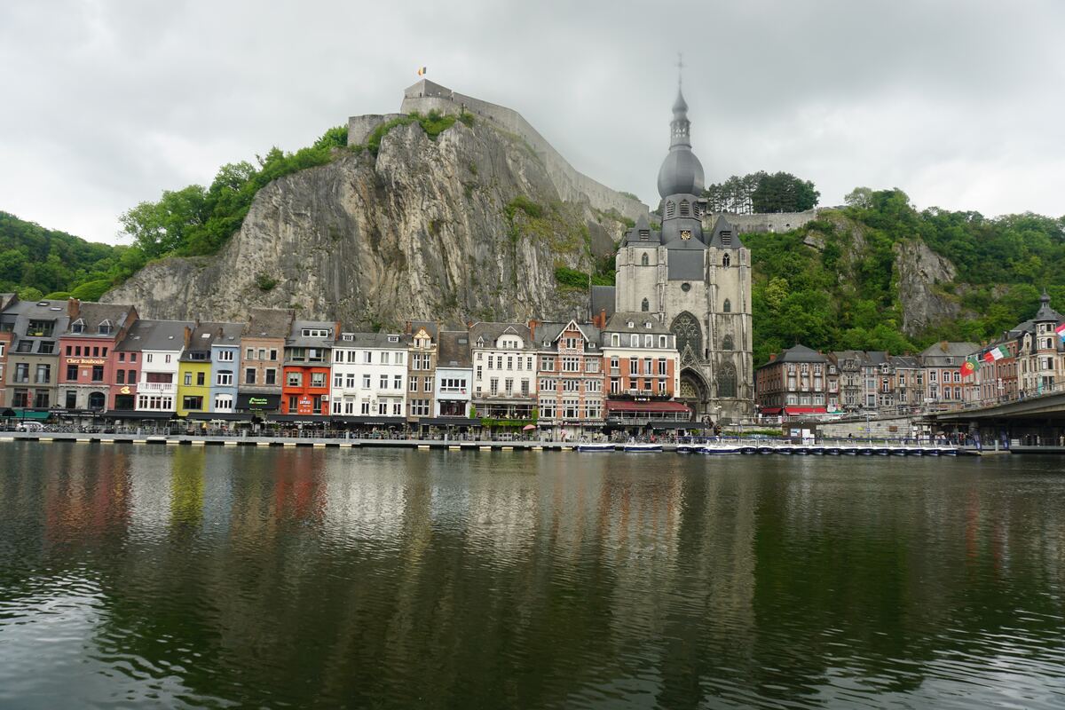 Een panorama van de stad Dinant, gezien vanaf het water, met de Maas op de voorgrond. Langs de oever staan kleurrijke gebouwen, waaronder restaurants en winkels. Op de achtergrond is een steile rotswand te zien met daarop de citadel van Dinant en de Collegiale kerk van Onze-Lieve-Vrouw.
