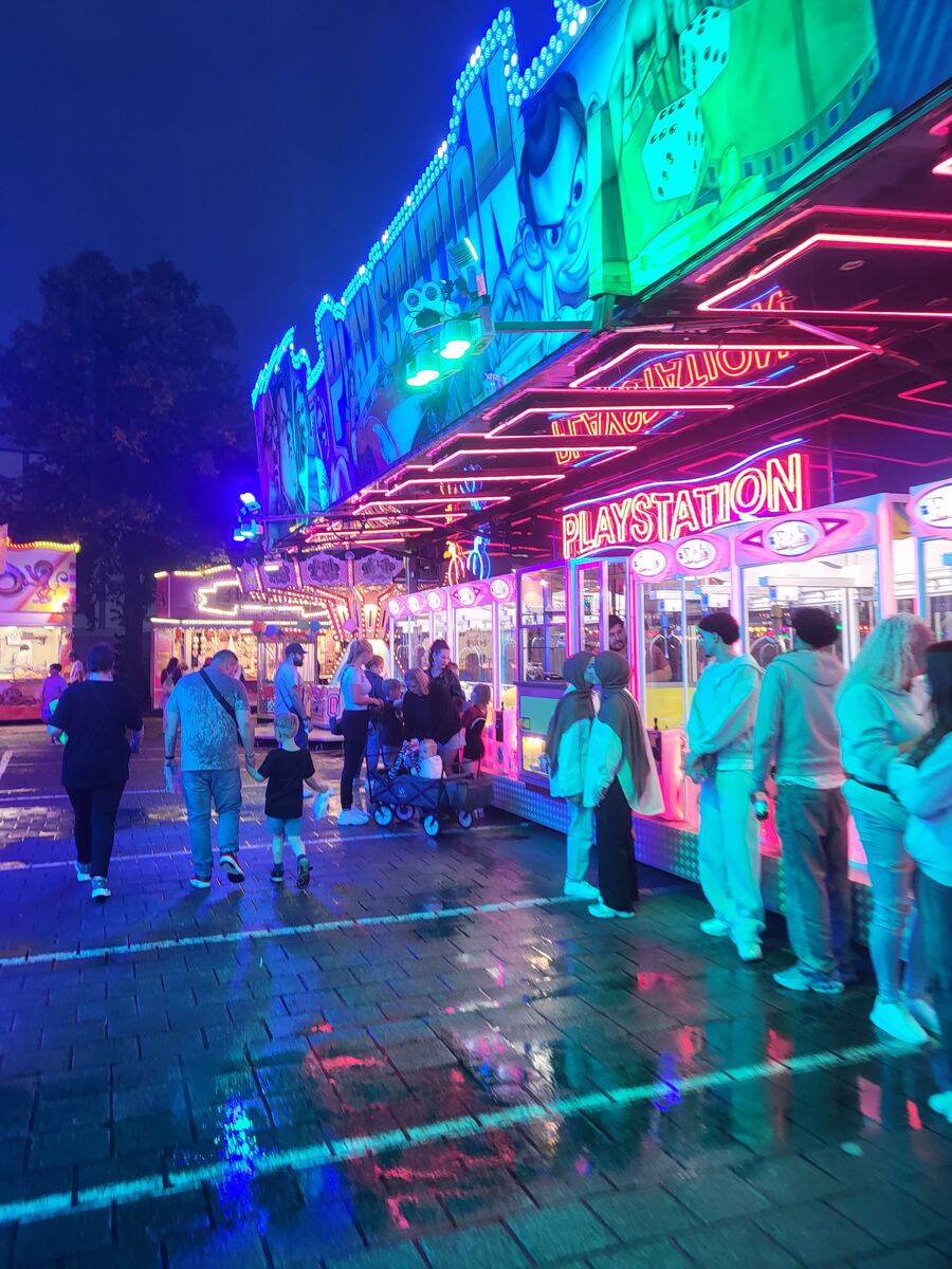 Een avondbeeld van een kermisattractie met veel neonverlichting in blauw, roze en groen. Er zijn mensen te zien die langs de attracties lopen, waaronder een spelletjeskraam met het woord 'PLAYSTATION' in neonletters. De grond is nat en reflecteert de felle lichten.