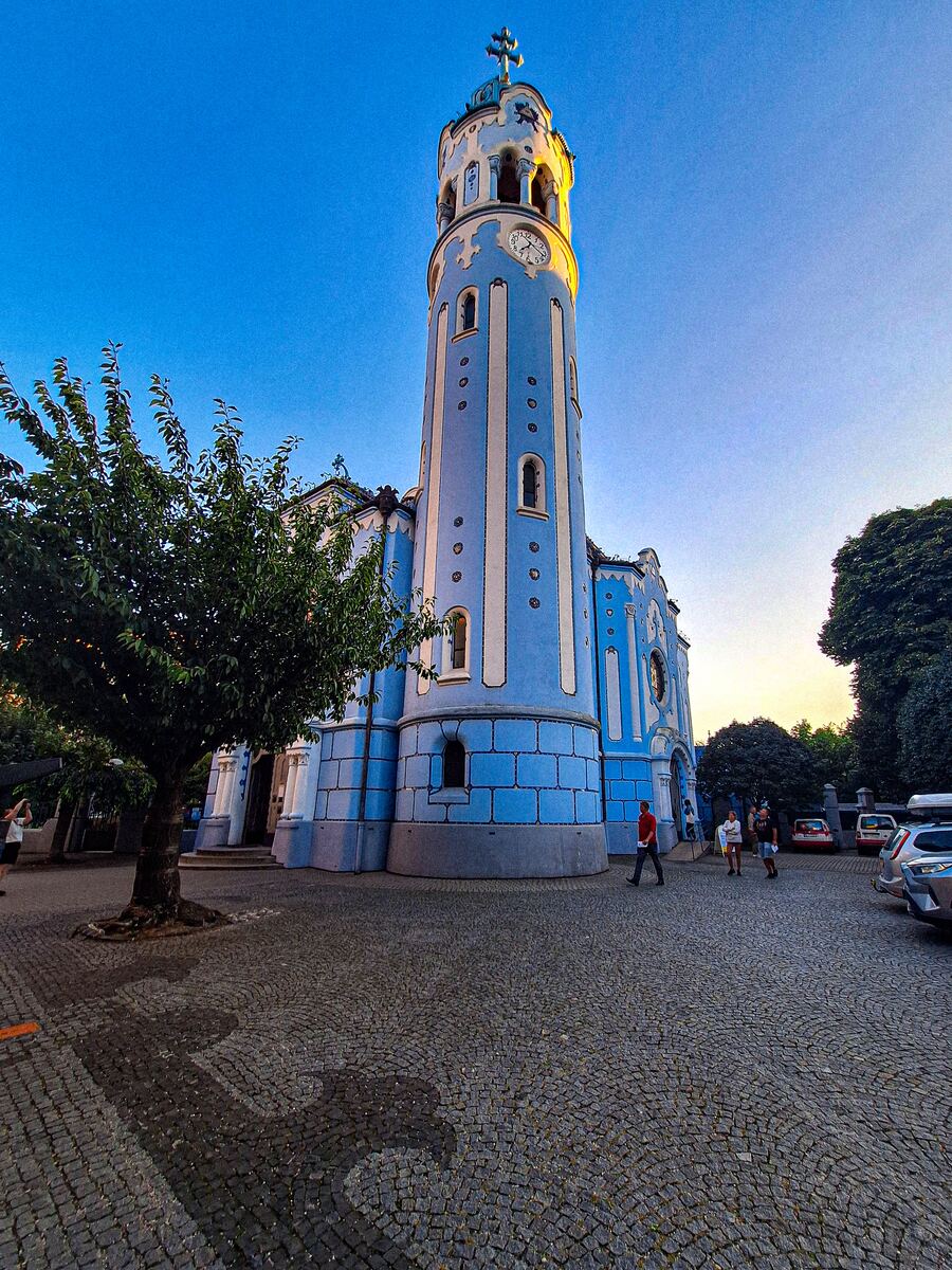 Een opvallende blauwe kerk met een hoge klokkentoren, omringd door bomen en een geplaveid plein, onder een helderblauwe lucht met een lichte zonsonderganggloed.