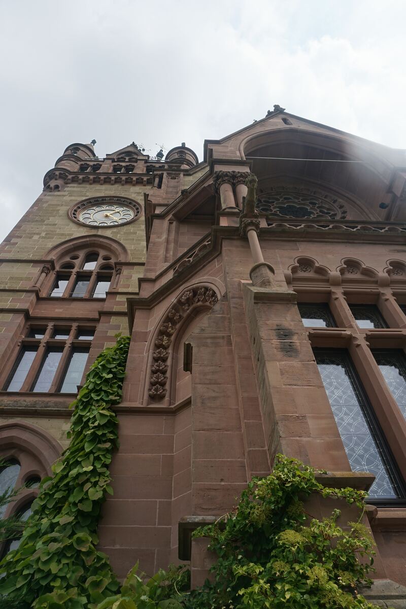 Een laag camerastandpunt van een groot, historisch gebouw met een klokkentoren, gemaakt van rode en lichtbruine stenen. Er is veel groen van klimplanten zichtbaar tegen de gevel en rond de ramen. De lucht is bewolkt.