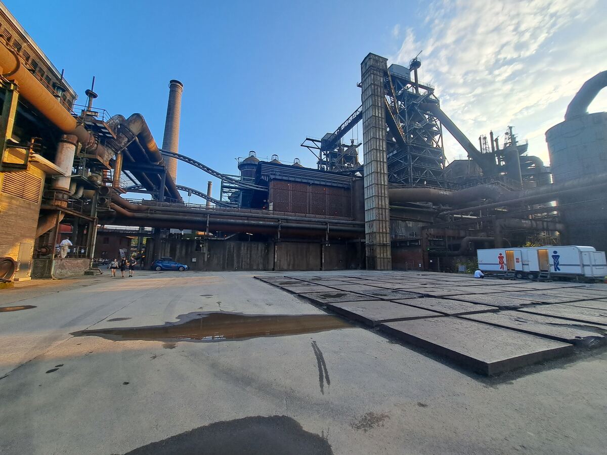 Een breed perspectief van een uitgestrekt industrieel complex met meerdere grote hoogovens, schoorstenen, pijpleidingen en stalen constructies onder een deels bewolkte blauwe avondlucht. De grond is een geplaveid terrein met enkele plassen. Rechts in beeld staat een witte trailer, mogelijk een mobiel toiletgebouw. Links op de achtergrond zijn enkele personen zichtbaar en een blauwe auto geparkeerd.