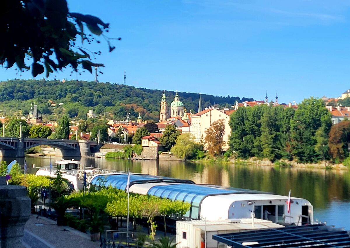 Een zonnig uitzicht over de Moldau in Praag, met een brug en gebouwen langs de rivier. Op de achtergrond is een groene heuvel met zendmasten te zien. Op de rivier liggen enkele rondvaartboten en aan de kade staan bomen en struiken.