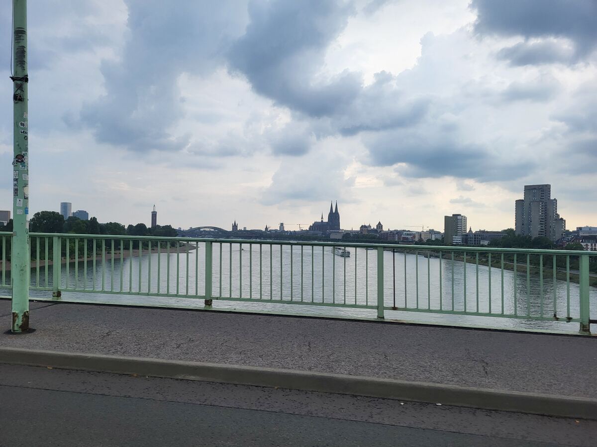 Een foto genomen vanaf een brug met een groen hekwerk, met uitzicht over een brede rivier. Aan de overkant van de rivier is de skyline van een stad te zien, met prominent de Dom van Keulen en andere gebouwen. De lucht is bewolkt.