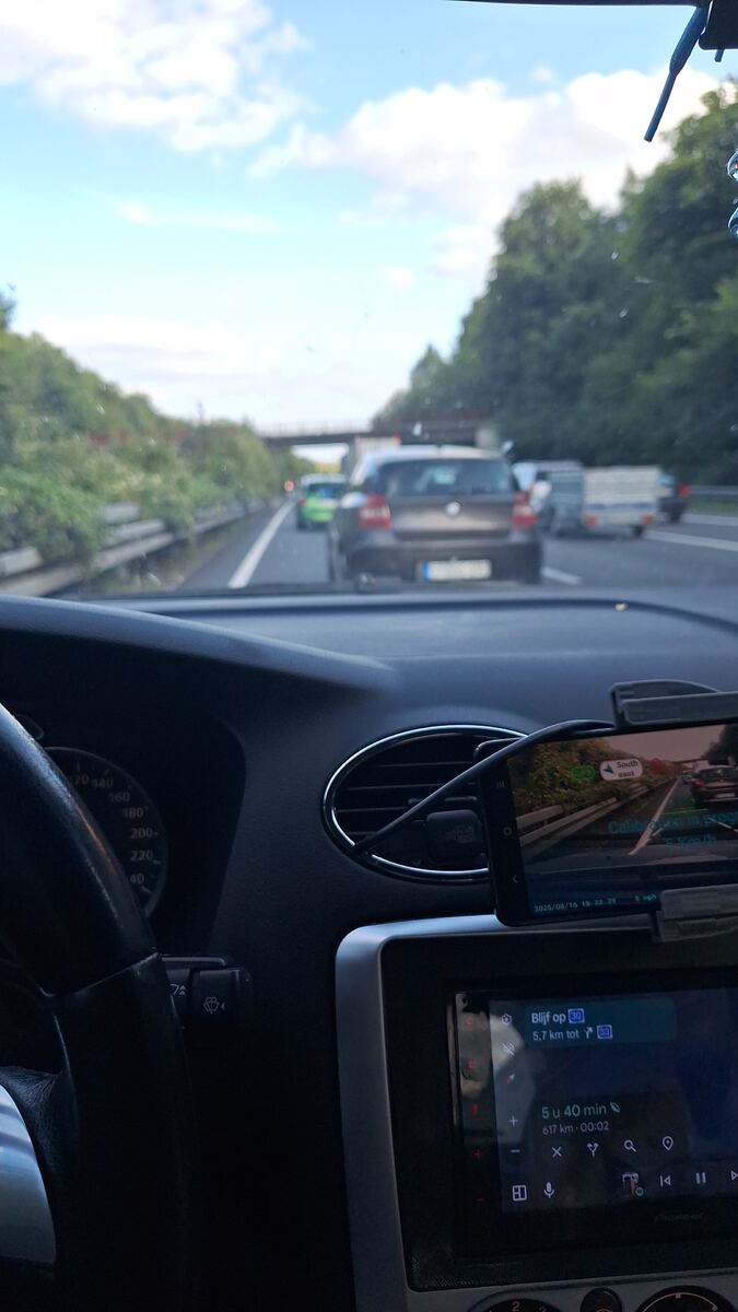 Een foto genomen vanuit de auto op een snelweg, met het dashboard en een navigatiescherm zichtbaar. Voor ons rijdt ander verkeer en in de verte is een viaduct te zien. De lucht is blauw met enkele wolken.