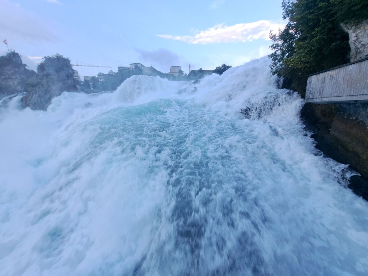 Een brede waterval vult het grootste deel van de voorgrond en het midden van de foto met kolkend, wit en turkoois water. Aan de rechterkant is een houten loopbrug te zien die langs de rotswand is gebouwd, deels overgroeid met groen blad. In de verte zijn boven de waterval enkele gebouwen en een kraan zichtbaar onder een lichtbewolkte avondlucht.