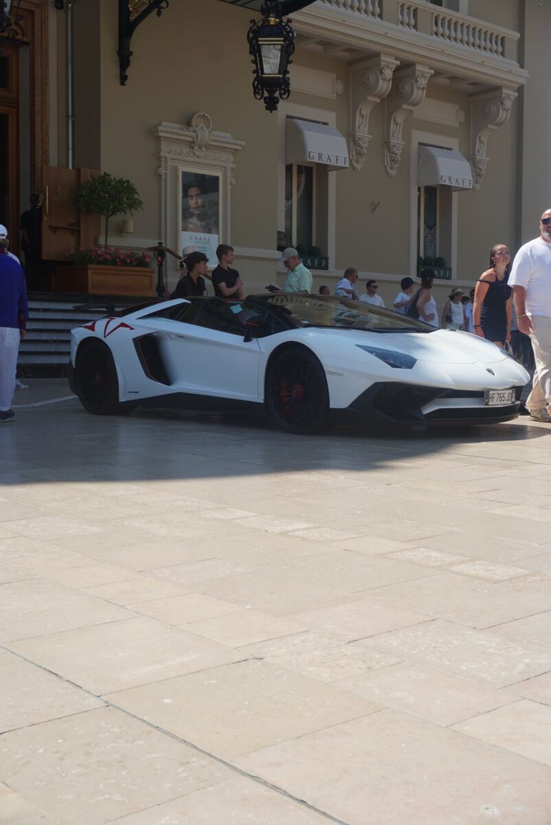 Een witte sportauto, waarschijnlijk een Lamborghini Aventador, met rode accenten staat geparkeerd op een plein van lichte tegels. Achter de auto bevindt zich een klassiek ogend gebouw met gedetailleerde architectuur en twee luifels met het opschrift 'GRAFF'. Er lopen en staan meerdere mensen rond de auto en het gebouw.