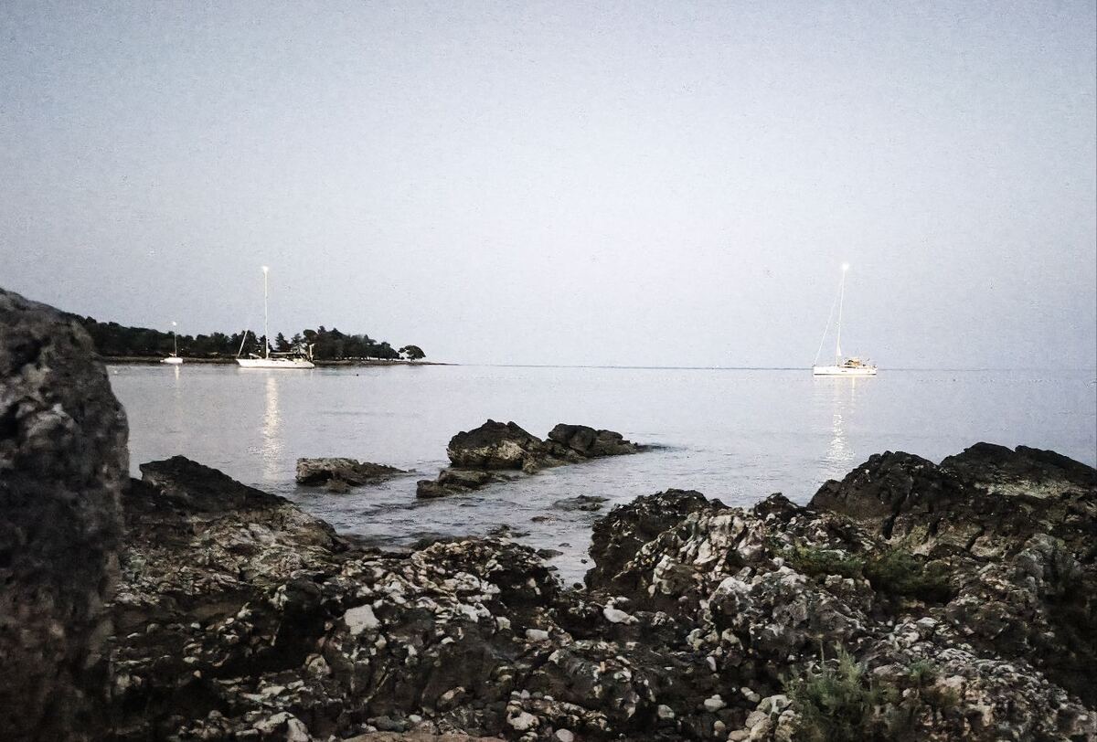 Een kustlandschap in de vroege ochtend of late avond, met op de voorgrond donkere rotsen en kalm water. Op de achtergrond zijn twee zeilboten zichtbaar, waarvan één met een verlichte mast. Aan de linkerkant is een beboste landtong te zien.