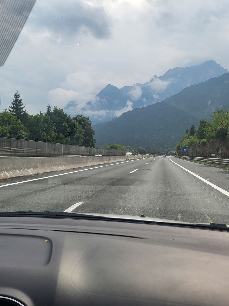Een foto genomen vanuit een auto op een snelweg, met op de achtergrond bergen die deels in de wolken liggen. De weg heeft twee rijstroken in elke richting, gescheiden door een betonnen barrière. Langs de weg staan bomen en een geluidswal. De lucht is bewolkt.