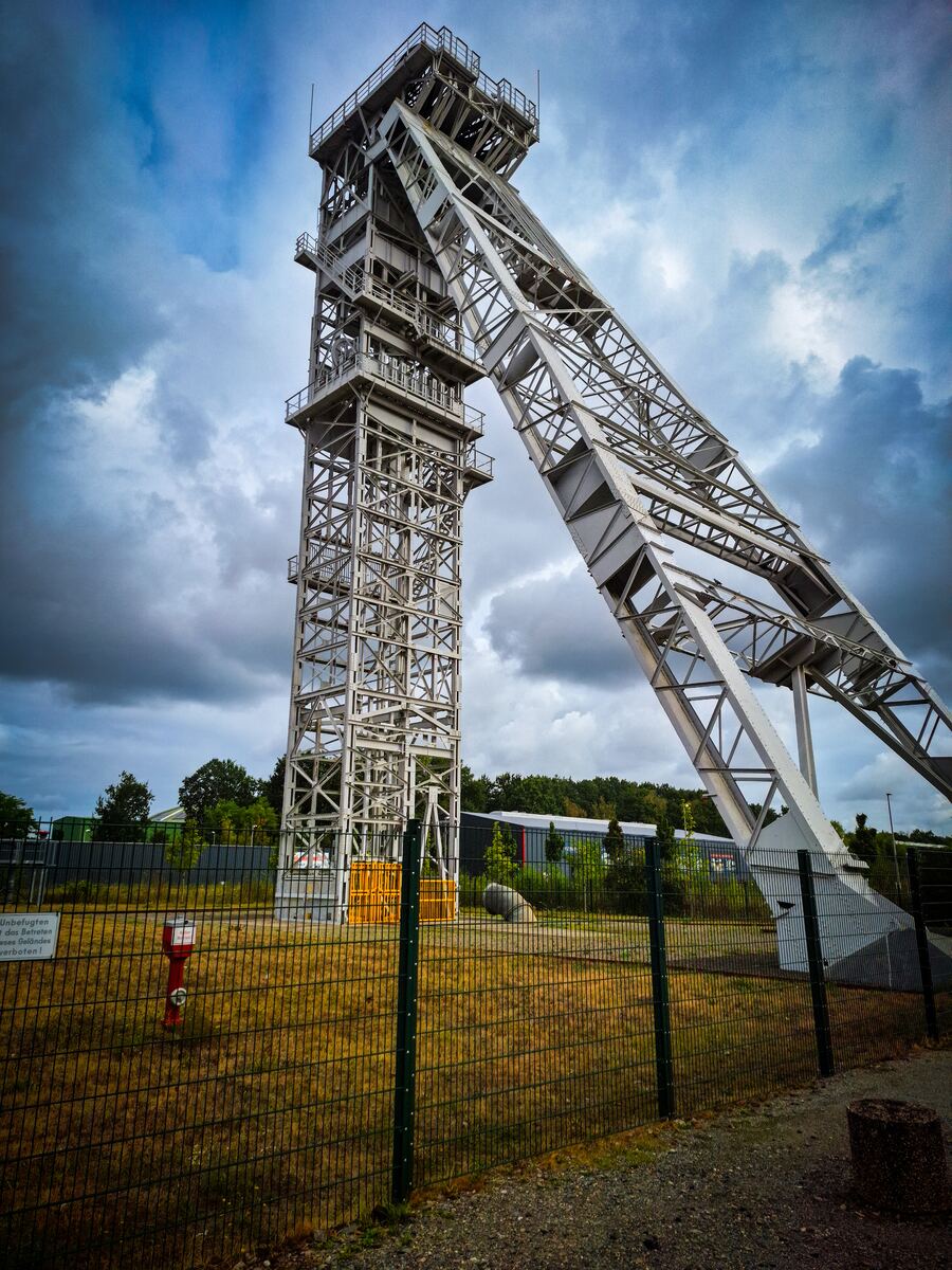Een hoge, stalen mijntoren met een schuine constructie ernaast, onder een bewolkte lucht. De toren staat achter een hek op een grasveld, met bomen en gebouwen op de achtergrond.