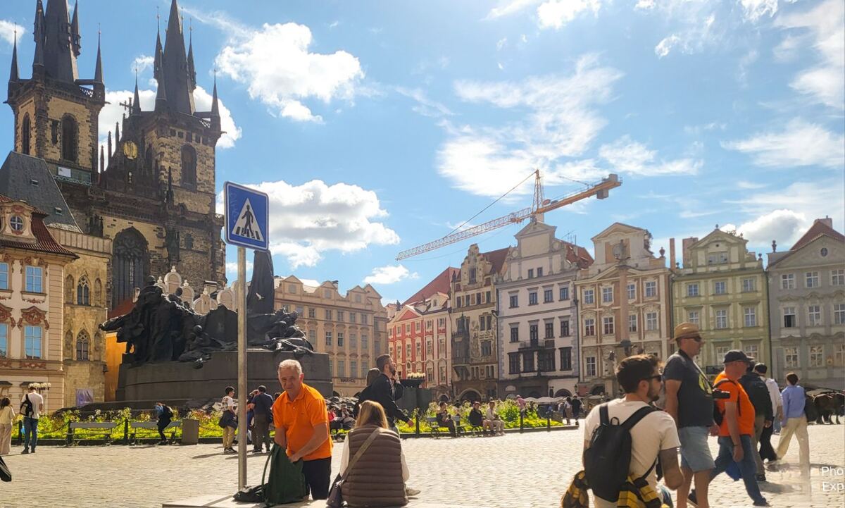Een zonnige dag op het Oude Stadsplein in Praag, met de Týnkerk en het Jan Hus-monument op de voorgrond. Er zijn veel mensen op het plein en op de achtergrond zijn historische gebouwen en een bouwlift te zien onder een helderblauwe lucht met witte wolken.