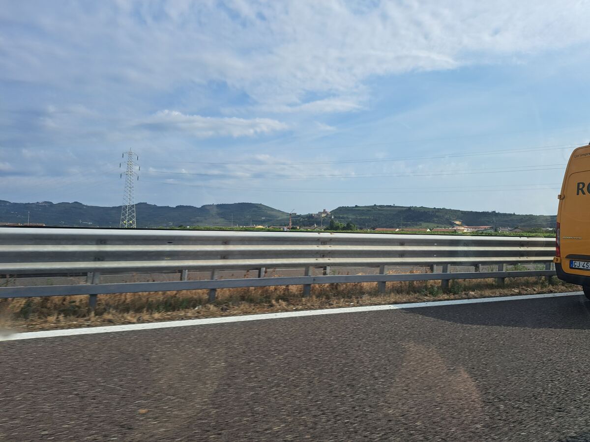 Een foto genomen vanuit een voertuig op een snelweg, met op de voorgrond de weg en een vangrail. Daarachter zijn glooiende, groene heuvels te zien, deels bedekt met wijngaarden. Op een van de heuvels staat een kasteel. Rechts is een deel van een gele bestelbus met een Italiaans kenteken zichtbaar. Een elektriciteitsmast staat in het landschap aan de linkerkant. De lucht is deels bewolkt.