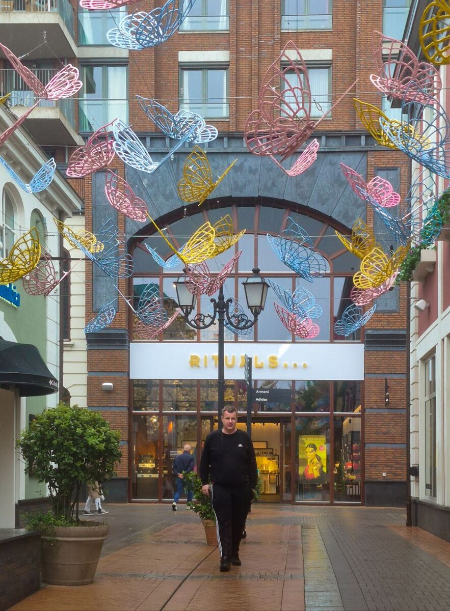Een man loopt over een bestrate winkelstraat onder een versiering van kleurrijke vlinders. Op de achtergrond is een Rituals-winkel te zien met daarboven een bakstenen gebouw. Links en rechts staan plantenbakken en andere winkels.