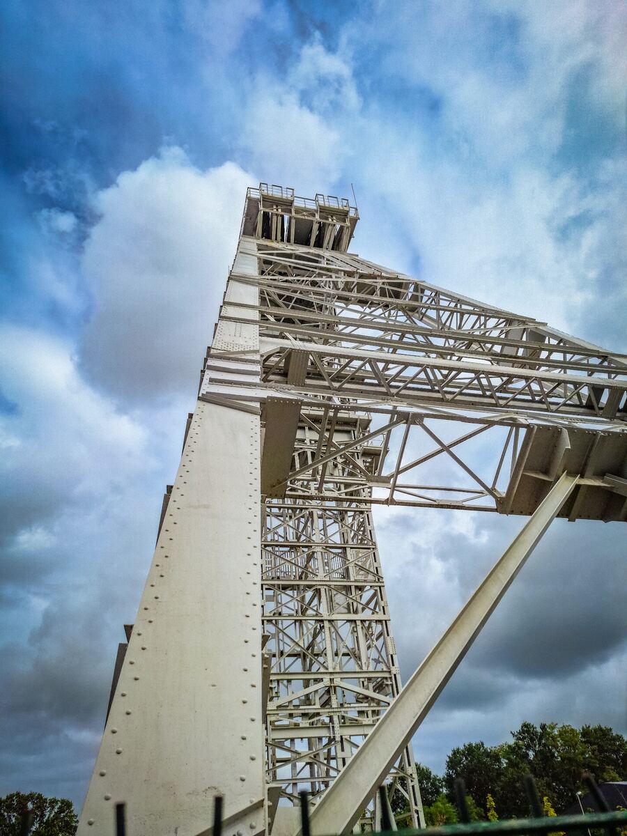 Een laag camerastandpunt toont een grote, witte metalen constructie die schuin omhoog loopt tegen een bewolkte blauwe lucht. De constructie bestaat uit een combinatie van massieve pilaren en een open vakwerkstructuur. Onderaan de foto zijn de toppen van groene bomen en een hek zichtbaar.