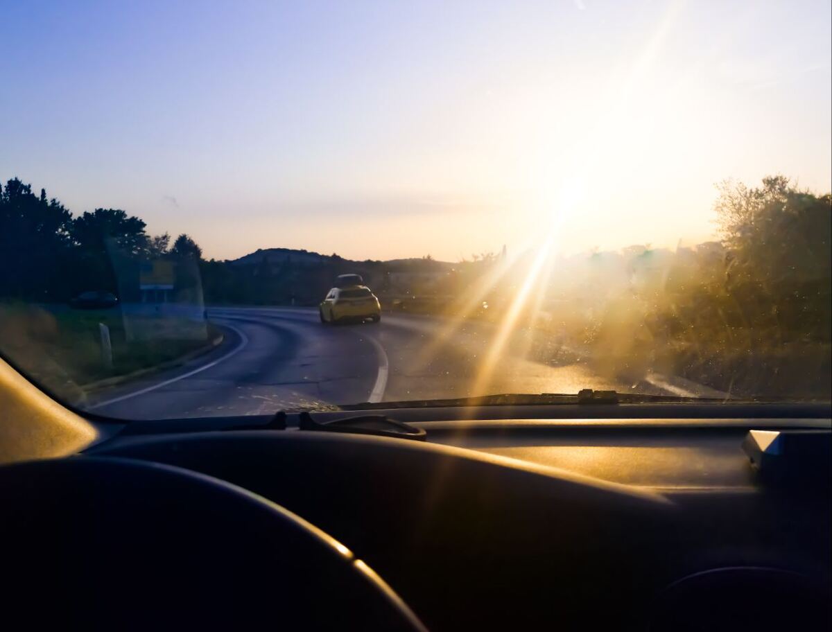 Een foto vanuit het interieur van een auto, genomen door de voorruit, met de zon laag aan de horizon die fel schijnt en reflecteert op de voorruit. Een gele auto met een dakkoffer rijdt voor ons uit op een bochtige weg. De omgeving bestaat uit heuvelachtig landschap met bomen en struiken, en de lucht is helderblauw met een oranje gloed van de opkomende zon.