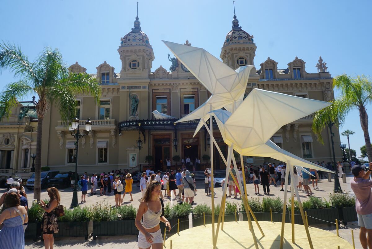 Een breed shot van de gevel van het Casino de Monte-Carlo in Monaco, met twee grote, witte, geometrische vogelsculpturen op de voorgrond. Voor het gebouw staan verschillende mensen en aan weerszijden zijn palmboomen zichtbaar. De lucht is helderblauw en de zon schijnt fel.