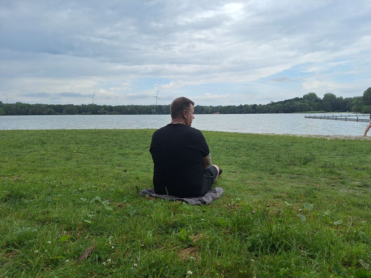 Een persoon zit op een handdoek in het gras met de rug naar de camera, kijkend over een meer. Aan de overkant van het water is een bosrand zichtbaar met enkele windmolens. De lucht is bewolkt.