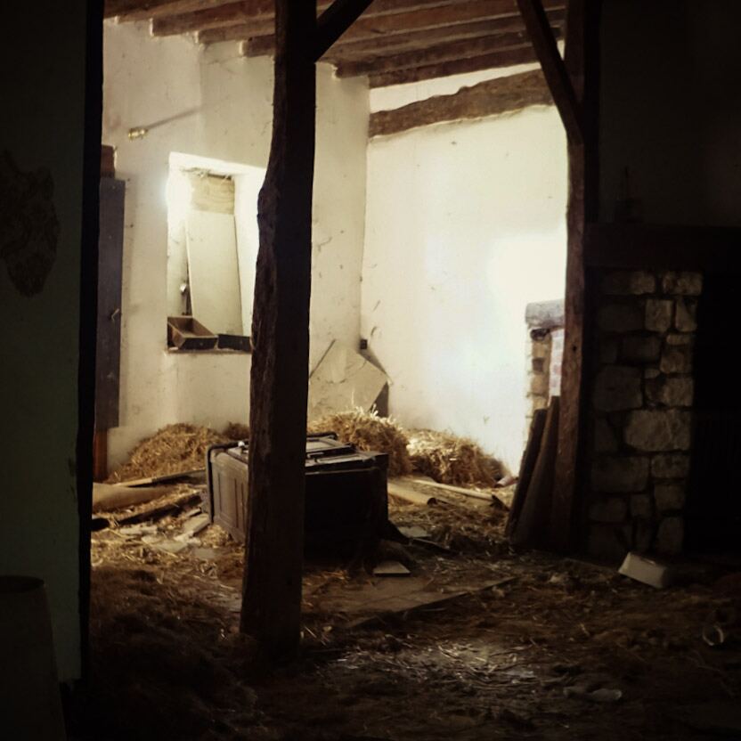 Een donkere, stoffige binnenruimte met houten balken, witte muren en stro op de grond. Er staat een oud meubelstuk in het midden en er is een raam zichtbaar waar licht doorheen komt. Rechts is een stenen muur of haard te zien.