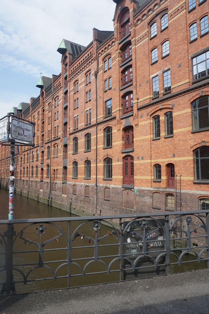 Een rij hoge, roodbruine bakstenen pakhuizen langs een gracht, gezien vanaf een brug. De gebouwen hebben meerdere verdiepingen met talrijke ramen, waarvan sommige met rode kozijnen, en kenmerkende gevels met groene dakaccenten. Het water in de gracht weerspiegelt de gebouwen. Op de voorgrond is een decoratieve metalen brugleuning zichtbaar, waaraan enkele hangslotjes en wat graffiti zijn bevestigd. Links staat een straatnaambord met de tekst 'Wandbereiter-Brücke'. De lucht is lichtbewolkt.