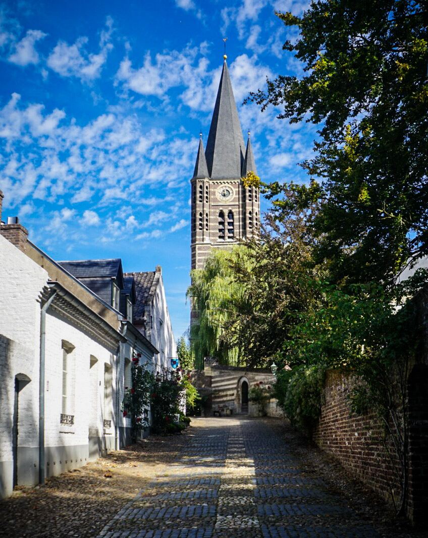 Een smal, geplaveid straatje met aan de linkerkant witgepleisterde huizen en aan de rechterkant een bakstenen muur met veel groen. Op de achtergrond domineert een hoge kerktoren met een spitse, donkere spits en een klok. De lucht is helderblauw met verspreide witte wolken.