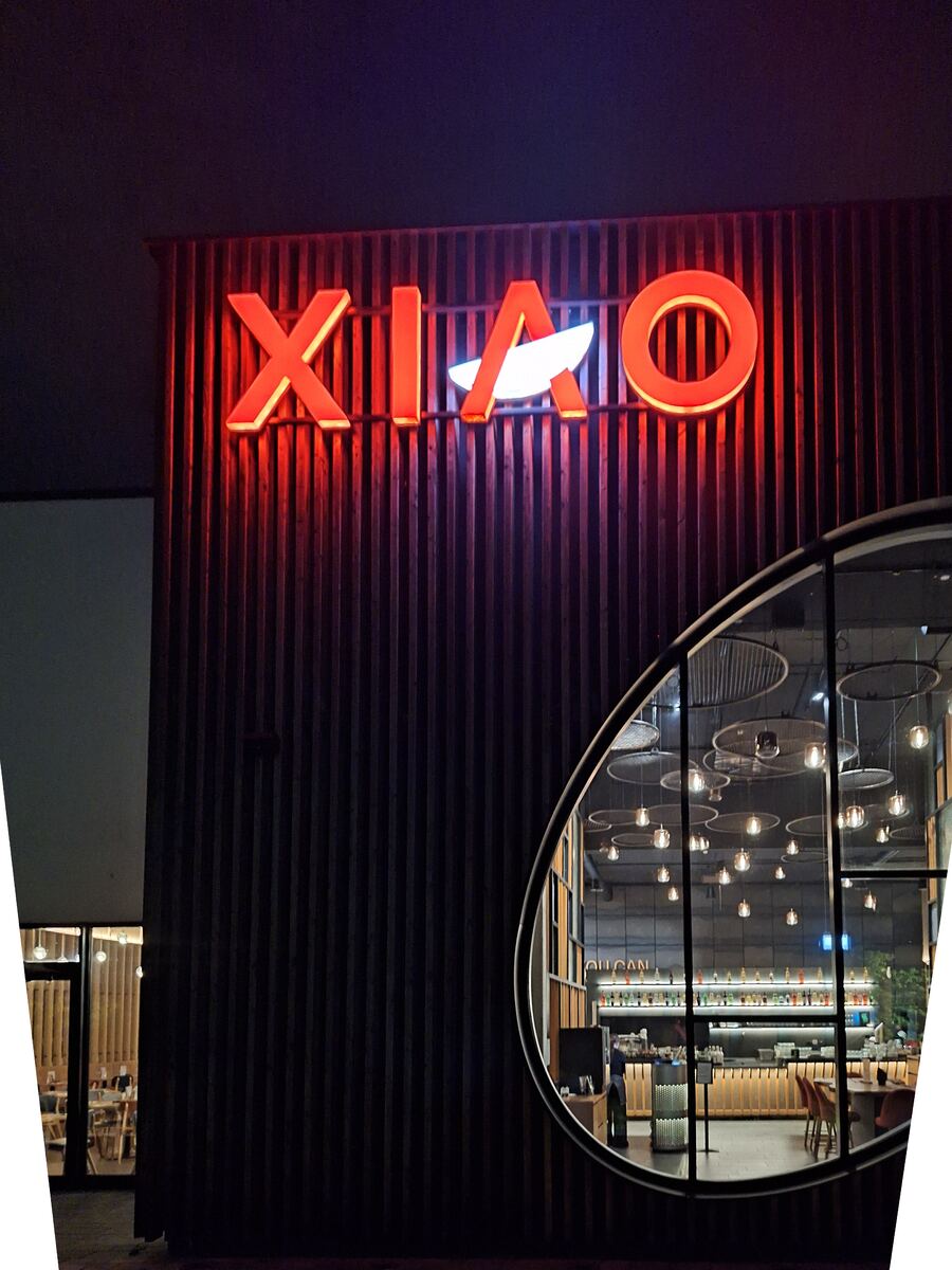 Een avondfoto van de buitenkant van een restaurant met een groot, rood neonlichtbord met de naam 'XIAO'. De gevel is bekleed met verticale houten latten. Een deel van het interieur is zichtbaar door een rond raam, met hangende lampen en een bar.