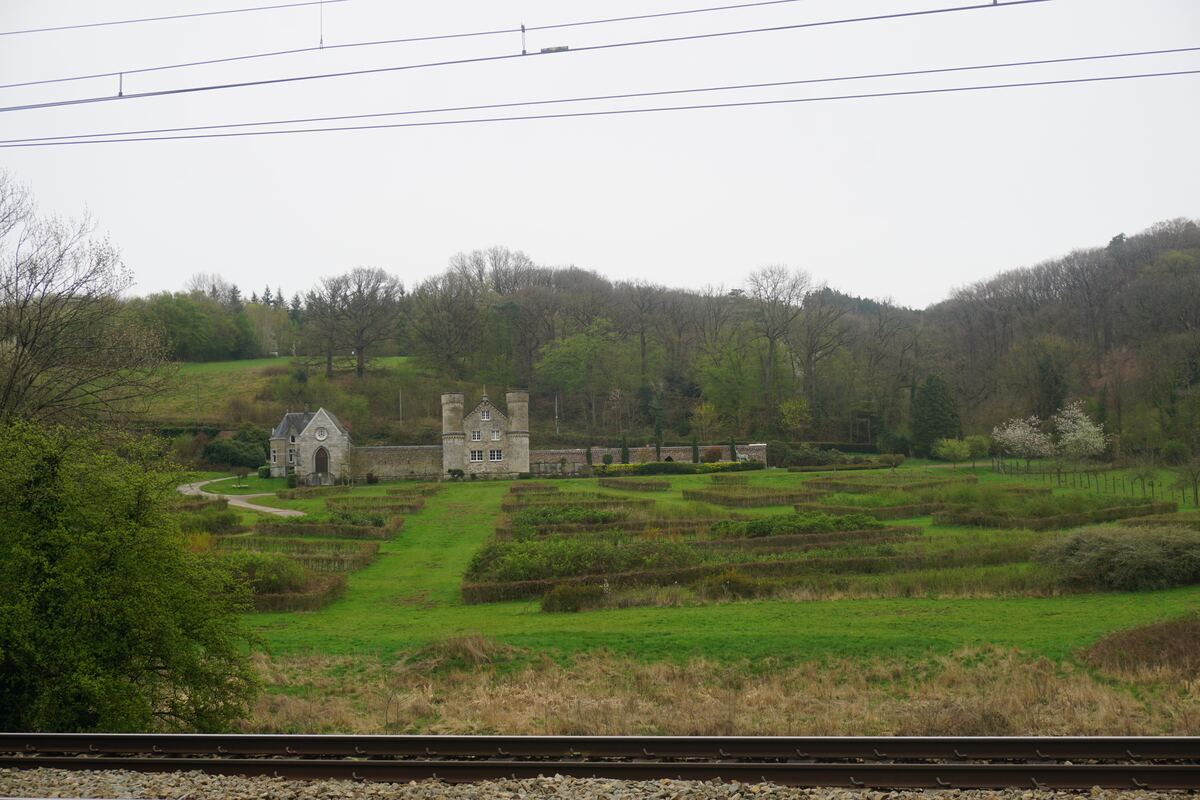 Een kasteelachtig gebouw met twee torens en een kleiner bijgebouw, omgeven door een muur en een trapsgewijs aangelegd groen landschap met heggen. Op de voorgrond zijn spoorrails zichtbaar en op de achtergrond een beboste heuvel onder een grijze lucht.