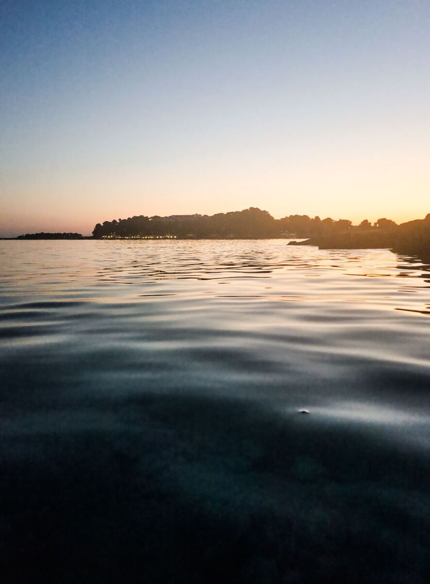 Een foto genomen vanaf het wateroppervlak met de horizon in de verte. De lucht toont een zonsondergang met kleuren die van blauw naar oranje en geel verlopen. Aan de horizon is een landtong met bomen en enkele verlichte gebouwen te zien. Het water op de voorgrond is donkerder en weerspiegelt de kleuren van de lucht.