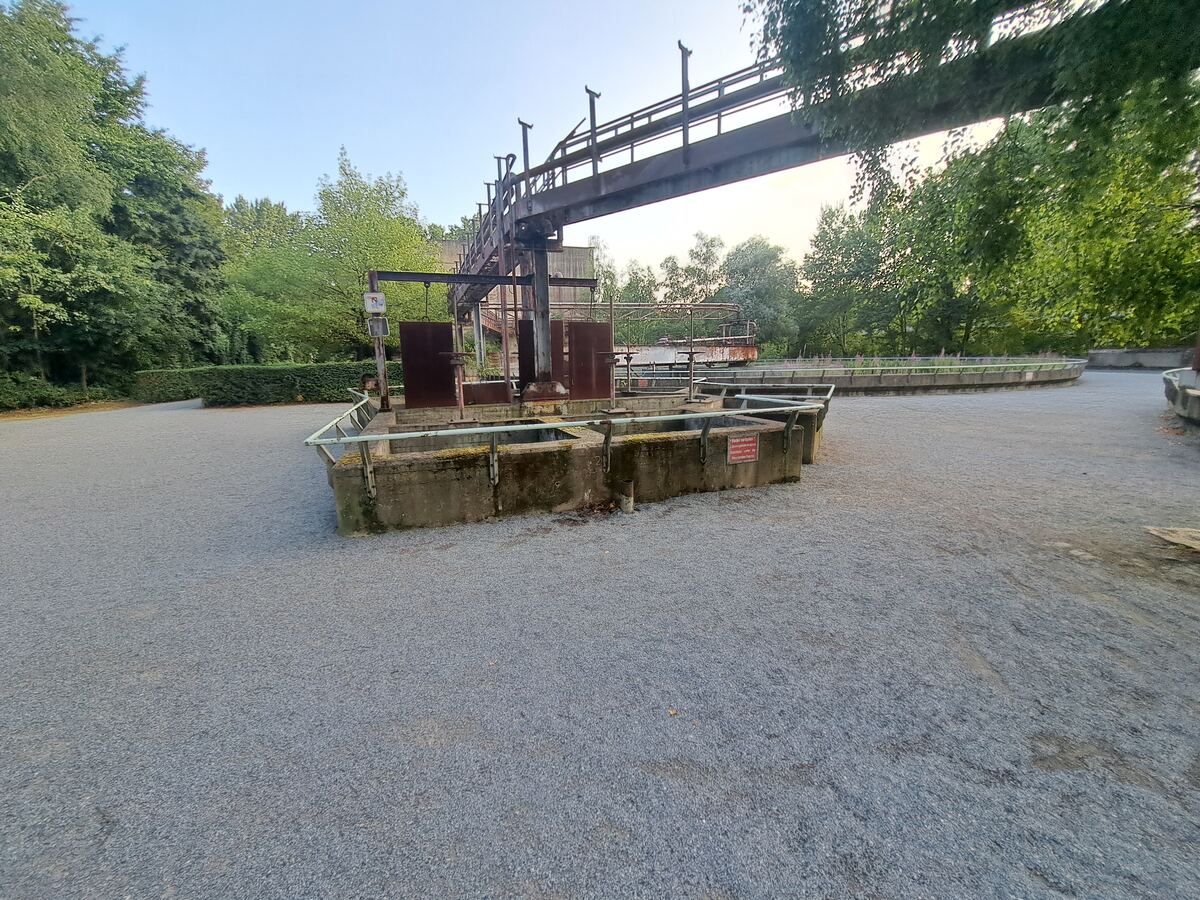 Een breed shot van een oud industrieel complex met betonnen bakken op de voorgrond en een grote, roestige metalen brugconstructie erboven. De grond is bedekt met grijs grind en rondom zijn veel groene bomen en struiken zichtbaar. De lucht is helderblauw.