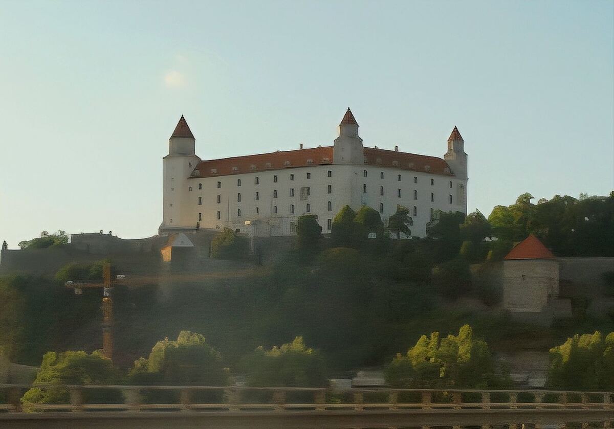 Een foto van de Burcht van Bratislava, een groot wit gebouw met vier hoektorens en rode daken, gelegen op een heuvel. De burcht is omgeven door groen en op de voorgrond is een deel van een brug of viaduct zichtbaar. De lucht is lichtblauw en helder.