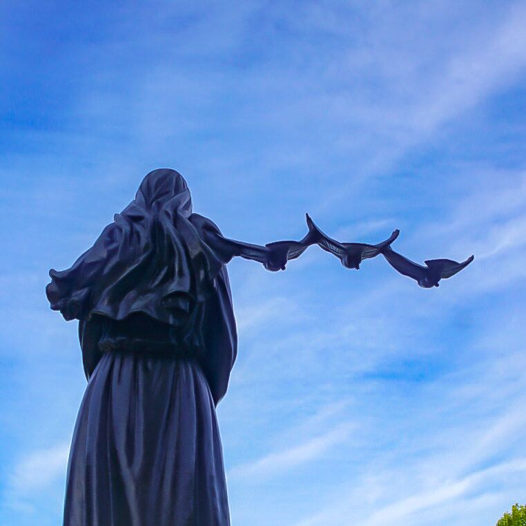 Een bronzen standbeeld van een figuur met een kap en gewaad, gezien van achteren, die drie vogels in vlucht lijkt los te laten tegen een helderblauwe lucht met enkele witte wolken.
