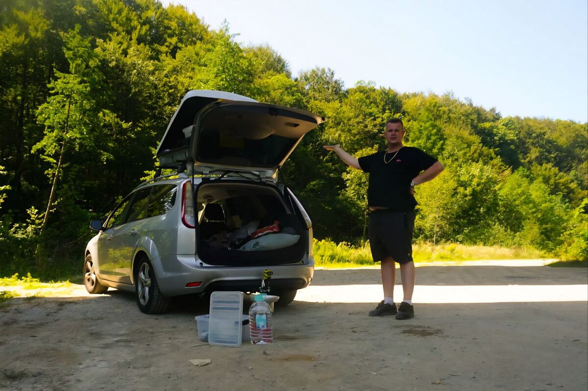 Een man staat naast een zilverkleurige Ford Focus stationwagen met open achterklep, geparkeerd op een onverharde weg. Achter de auto is een dichtbegroeid bos te zien. Op de grond naast de auto staan enkele plastic bakken en een fles water.