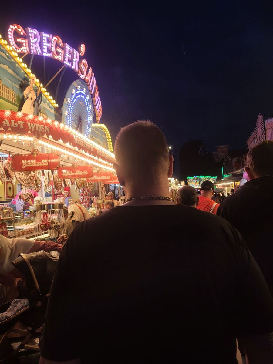 Een foto genomen vanaf de rug van een persoon, kijkend naar een verlichte kermiskraam met de tekst 'GREGER'S' en 'ALT WIEN'. De kraam verkoopt snoepgoed zoals 'Zuckerwatte' en 'Lebkuchenherzen'. Op de achtergrond is een ander kermisattractie te zien met blauwe verlichting. De lucht is donker, wat duidt op de avond. Er zijn meerdere mensen zichtbaar op de kermis.