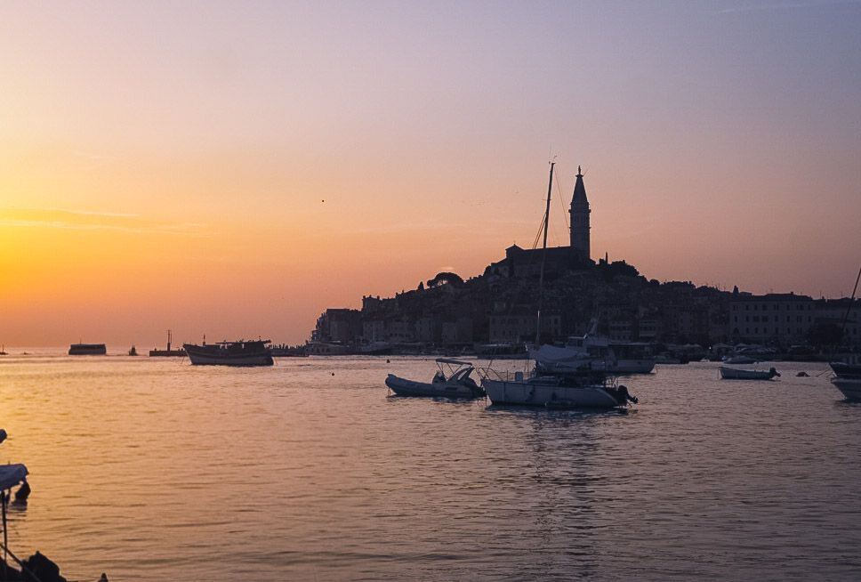 Een avondopname van de kustlijn van Rovinj, Kroatië, met de stad en de klokkentoren van de Sint-Eufemiakerk als silhouet tegen een oranje-paarse zonsondergang. Op het water liggen meerdere boten en jachten.