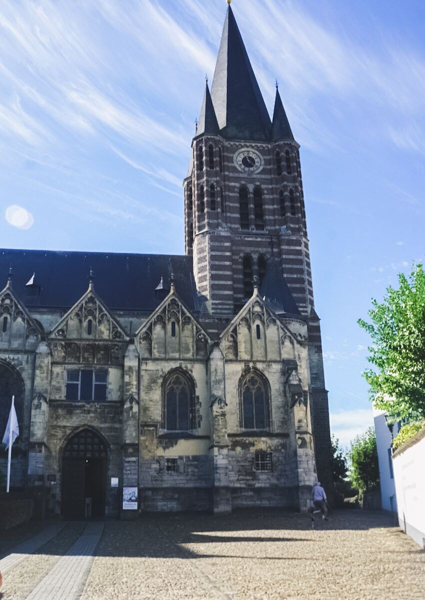 Een grote, oude kerk met een hoge toren en een klok, gefotografeerd op een zonnige dag met een blauwe lucht en enkele wolken. De kerk heeft een mix van baksteen en lichte steen, met gedetailleerde architectuur. Op de voorgrond is een geplaveid plein te zien en rechts staat een boom en een wit gebouw.