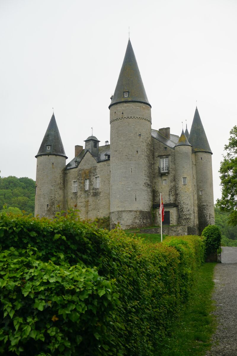 Een stenen kasteel met meerdere puntige torens, omgeven door groene heggen en bomen, onder een bewolkte hemel. Een rood-wit-rode vlag wappert bij de ingang.