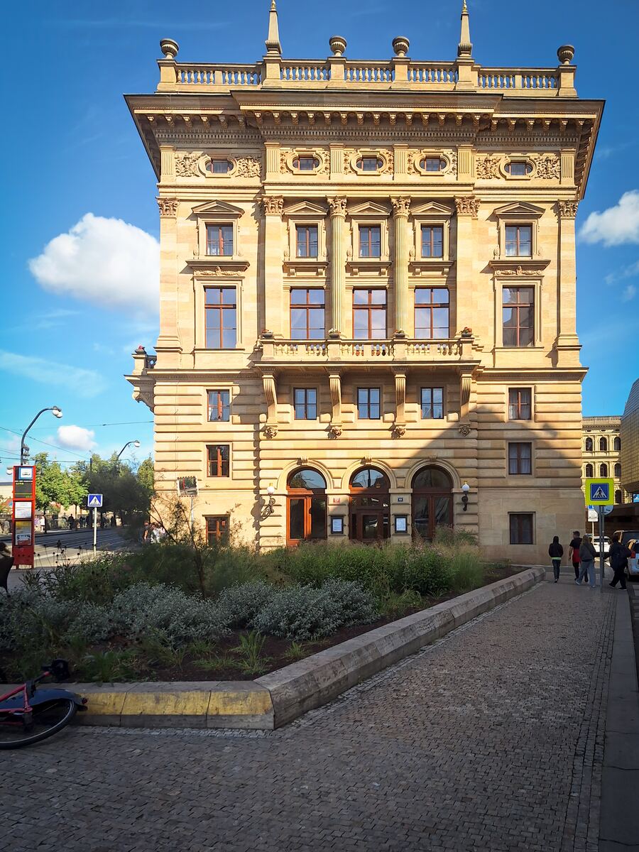 Een imposant, lichtgeel gebouw met veel decoratieve details, zuilen en ramen staat centraal in de foto. Op de voorgrond is een groenperk met lage beplanting te zien, en rechts loopt een geplaveid pad waar enkele mensen lopen. Links van het gebouw is een straat met verkeersborden en bomen zichtbaar onder een helderblauwe lucht met enkele wolken.